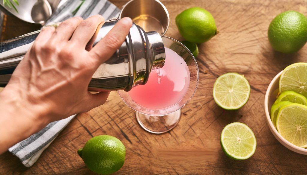 Top view of a hand straining a Cosmo cocktail from a cocktail shaker into a glass on top of a wooden kitchen counter