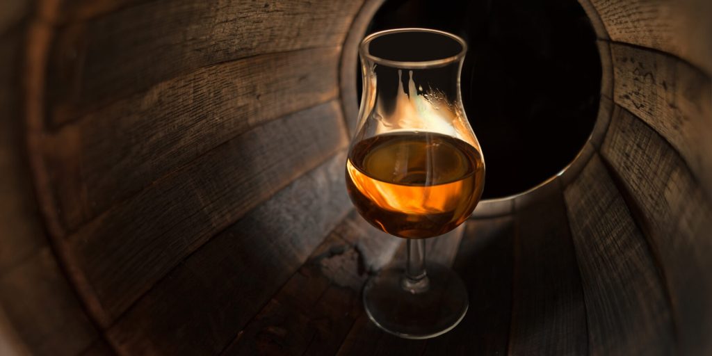 Tulip-shaped brandy glass inside a large wooden barrel, filled with amber brandy, photo by razoomanetu/Adobe Stock