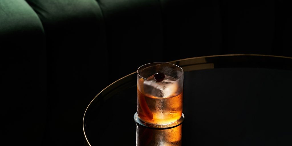 Classic bourbon cocktail served on the rocks with an orange peel and cherry garnish, set on a dark, glossy table in a moody, upscale setting.