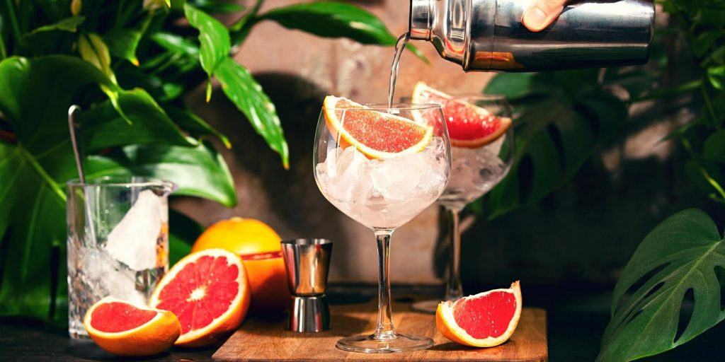 Close up of two Mezcal Paloma Cocktails being poured into large wine glasses, garnished with fresh slices of blood orange