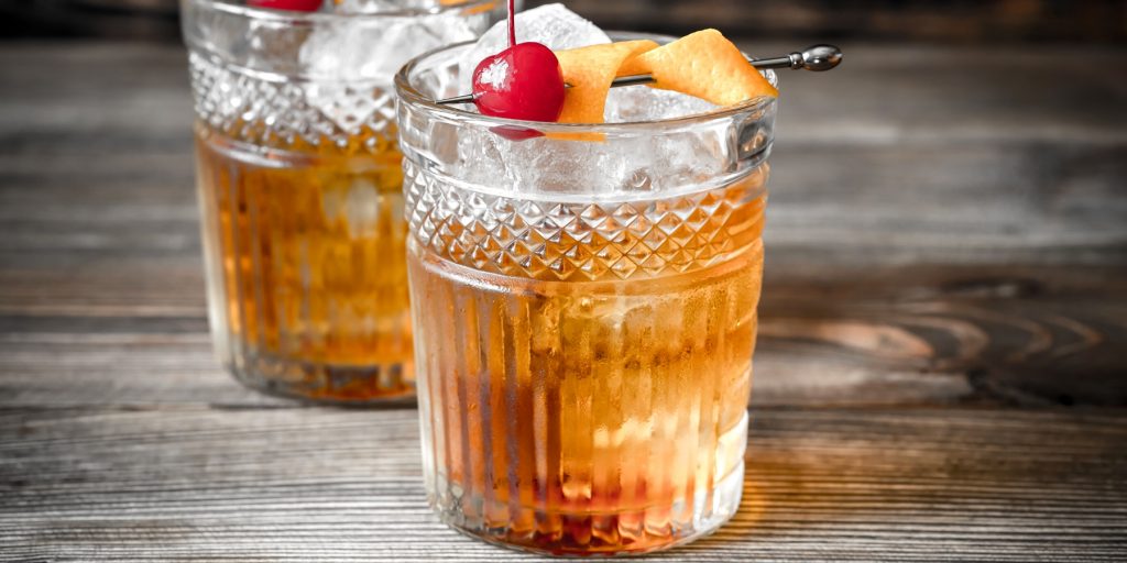 Close-up of two Scotch Old Fashioned cocktails in intricate glass tumblers garnished with cherries and orange twists on cocktail picks set on a grey surface