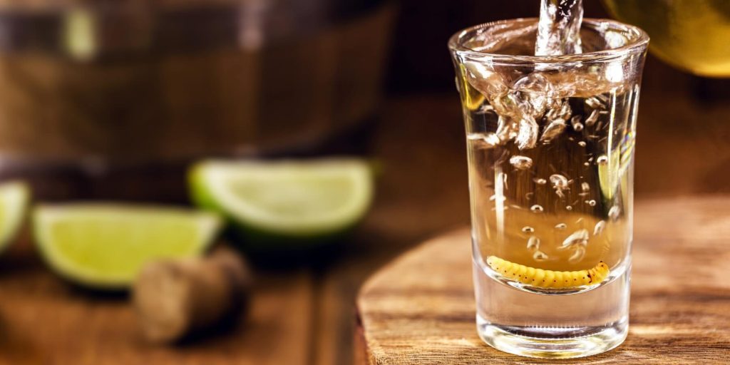 Pouring mezcal into a shot glass with a gusano inside, surrounded by lime wedges, photo by RHJ/Adobe Stock 