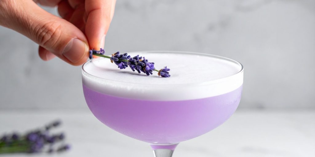 lose-up of a hand placing a lavender sprig on a foamy Lavender Gin Fizz cocktail in a coupe glass.