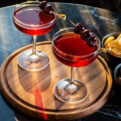 Two Manhattan cocktails with cherry garnishes on a wooden tray set on a marble table