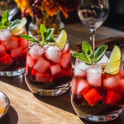 Three glasses of Thanksgiving batch cocktail Sparkling Pomegranate Punch, filled with ice and garnished with lime wedges and mint sprigs. The red drinks are displayed on a wooden table with place settings and fall florals.