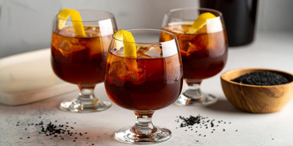 Three rounded glasses of the Boulevardier cocktail, a deep amber drink served over ice and garnished with a twist of lemon. Black sesame seeds are scattered on the table, hinting at the cocktail's unique twist.