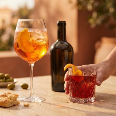 Two premixed cocktails — Aperol Spritz and Negroni — sharp on a travertine surface, one hand reaching in, golden-hour light.