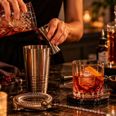 Close-up of a person measuring an amber cocktail into a shaker with a jigger, surrounded by bar tools, ice, citrus and cocktail garnishes.