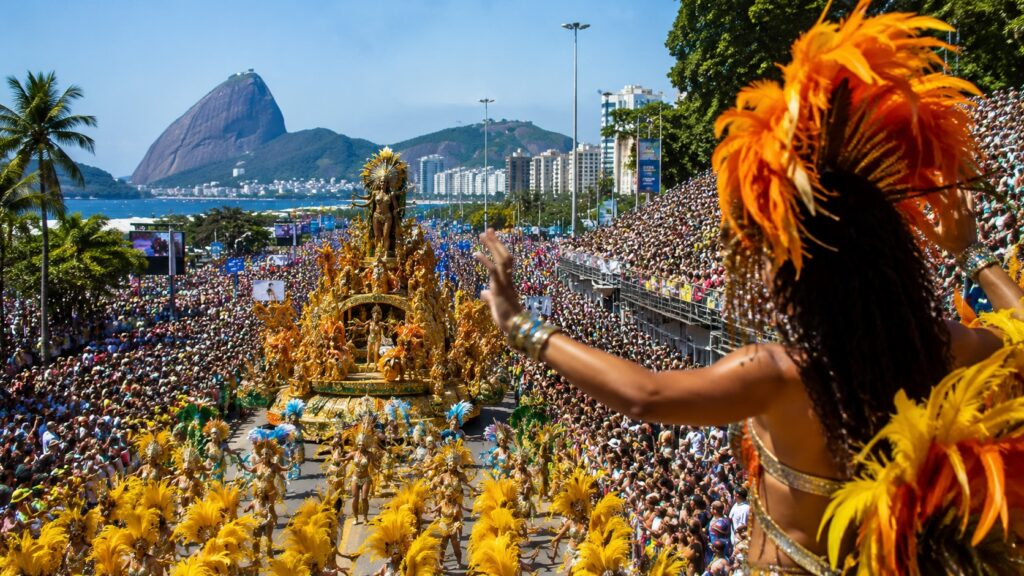 A panoramic view of a vibrant Brazilian Carnival parade with elaborate samba dancers in feathered costumes, colorful floats, and thousands of spectators lining the avenue.