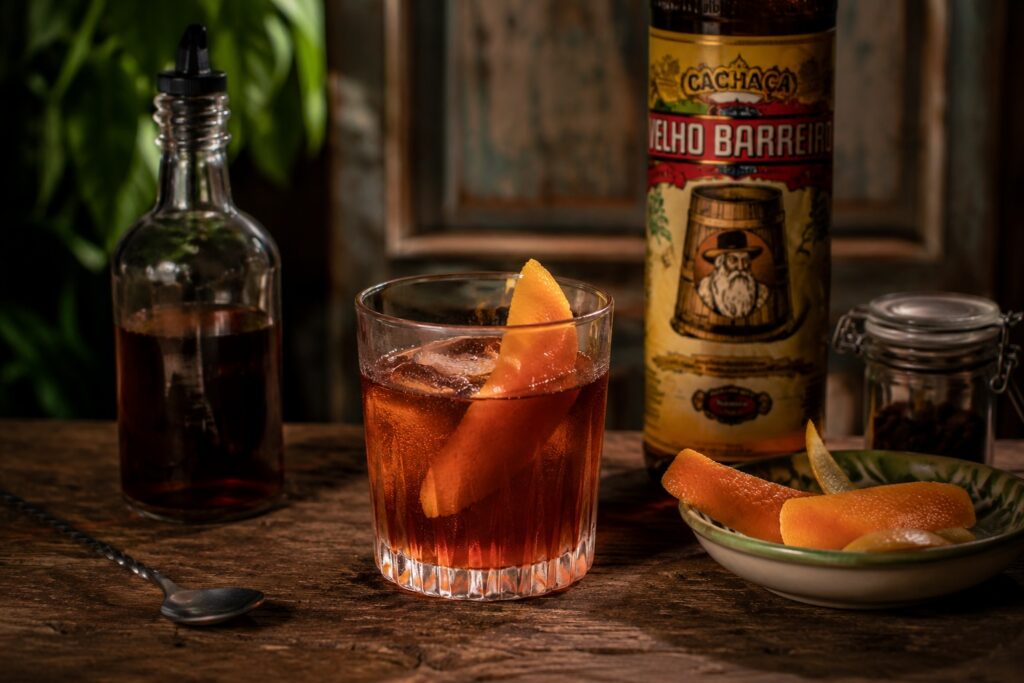 A Rabo de Galo cocktail served over ice in a rocks glass with an orange peel garnish on a rustic wooden bar.